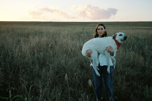 Ken Pomeroy stands in a field at sunset, holding a white dog with a red bandana around its neck. Pomeroy is looking at the camera and smiling slightly, wearing bluejeans and a white top.